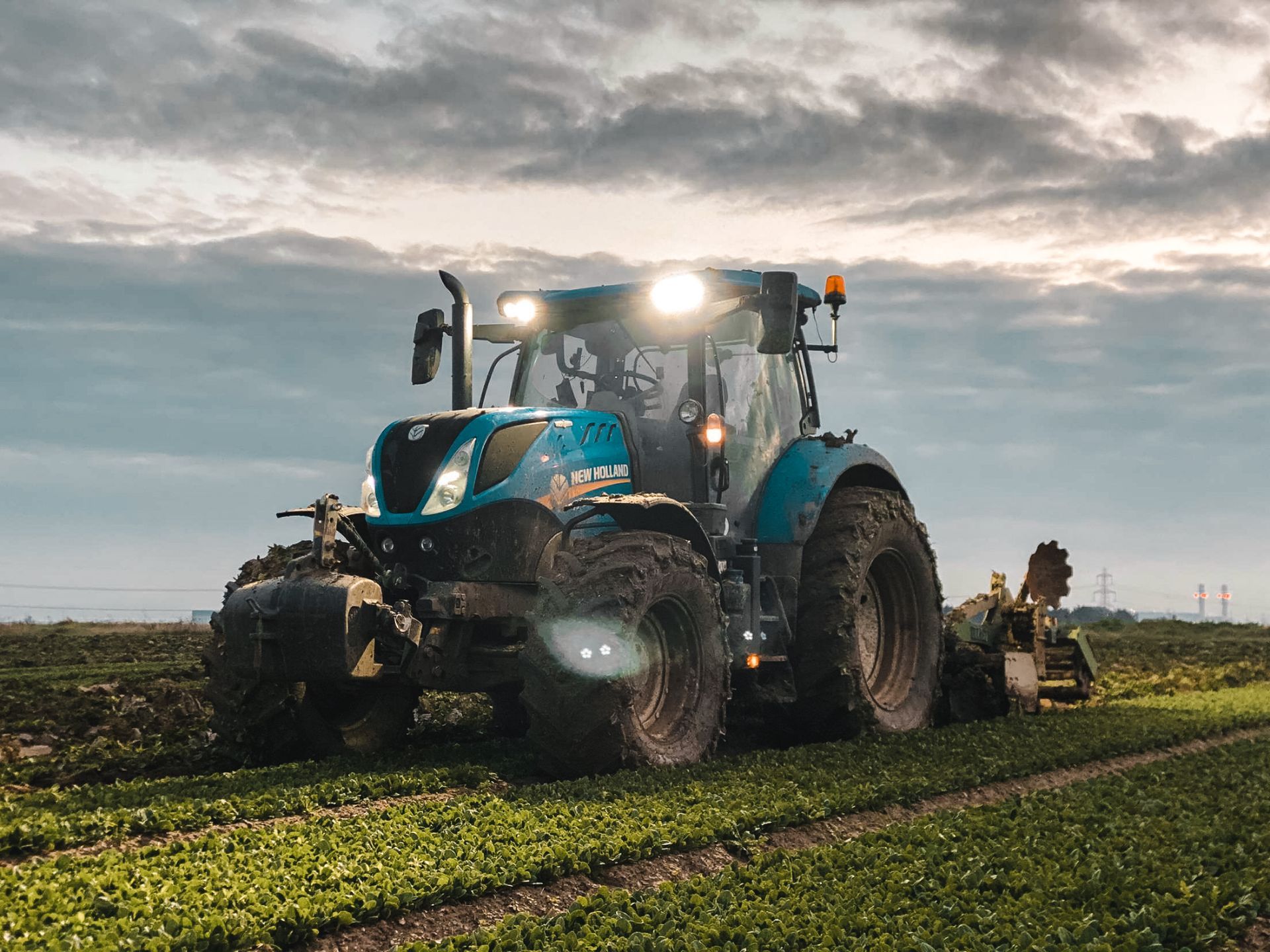Muddy blue tractor with its lights on, working a field under a cloudy sky